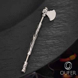 Puede incluir: Una joya para el cuerpo de color plateado con forma de hacha pequeña. La cabeza del hacha es de plata lisa y pulida, y el mango tiene un diseño texturizado y ondulado. La joya está sobre un fondo oscuro.