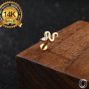 Può includere: Un piercing per il corpo a forma di serpente color oro con una gemma trasparente taglio marquise. Il gioiello è contrassegnato con "14K GOLD" ed è esposto su una superficie di legno. È visibile anche il marchio "OUFER BODY JEWELRY".