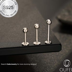 May include: Three silver lip piercing studs with ball tops, displayed on a dark surface. The studs are of varying lengths. The image includes a silver circular logo with the text "STERLING SILVER S925 STERLING SILVER". The text "Search OuferJewelry for more stunning designs!" is also visible.