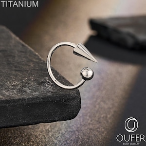 May include: A silver-colored titanium circular body jewelry ring featuring a pointed cone-shaped end and a smooth spherical ball end. The text TITANIUM is visible in the upper left corner, and the OUFER BODY JEWELRY logo is in the bottom right.