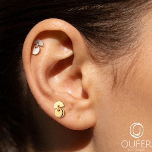 May include: A silver-toned semi-circle stud earring with a dangling circular charm in the upper ear cartilage and a matching gold-toned semi-circle stud earring with a dangling circular charm in the earlobe, featuring the text OUFER BODY JEWELRY.