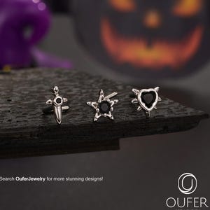 Op de afbeelding: Drie zilveren lichaamsjuwelen met zwarte edelstenen worden getoond op een donkere ondergrond. De stukken omvatten een dolk, een ster en een hartvorm. De achtergrond toont een vage Halloween-afbeelding. De tekst "Search OuferJewelry for more stunning designs!" is zichtbaar.