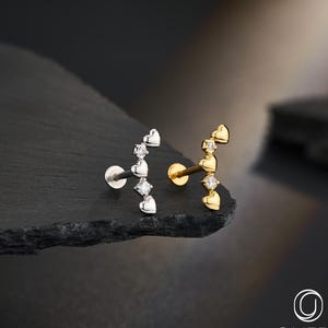 May include: Two pieces of body jewellery, one silver and one gold, each with a curved design of hearts and diamonds. The jewellery is displayed on a dark, textured surface. The brand name "OUFER BODY JEWELRY" is visible.