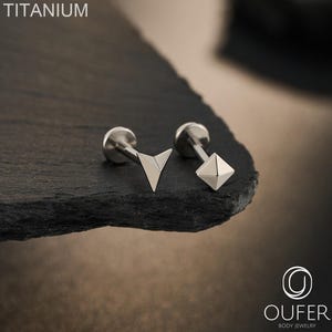 Può includere: Due piercing labret in titanio color argento con base piatta. Un piercing presenta una parte superiore a forma di freccia triangolare, mentre l'altro ha una parte superiore a forma di piramide a quattro lati. Il testo TITANIUM e OUFER BODY JEWELRY è visibile.