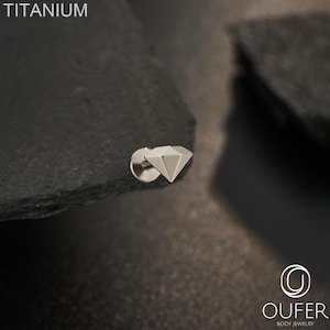 May include: A silver-colored titanium body jewelry stud featuring a faceted diamond-shaped top. The text TITANIUM is visible in the upper left corner, and the logo OUFER BODY JEWELRY is printed in the lower right corner of the image.