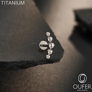 May include: A silver-colored titanium body jewelry piece featuring a curved bar with five graduated spherical beads arranged in a vertical arc. The text TITANIUM is visible in the upper left corner, and the OUFER BODY JEWELRY logo is in the bottom right.