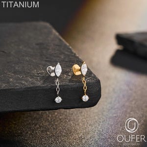 May include: Two titanium body jewellery pieces, one silver-toned and one gold-toned, each featuring a marquise-cut clear stone stud connected by a short chain to a small round clear stone dangle. The text TITANIUM and OUFER are visible.