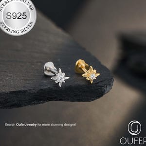以下が含まれることがあります： 星型のボディジュエリースタッド2つ。1つはシルバー、もう1つはゴールドで、それぞれ中央に宝石が付いています。シルバーのスタッドには、小さな宝石のクラスターがあります。「STERLING SILVER S925 STERLING SILVER」と「Search OuferJewelry for more stunning designs!」の文字が含まれています。
