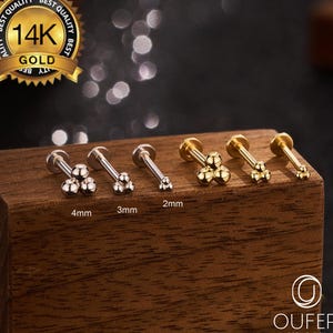 Puede incluir: Una colección de pendientes de joyería corporal dorados y plateados. Cada pendiente presenta un grupo de pequeñas esferas. Las tallas están etiquetadas como 2 mm, 3 mm y 4 mm. Un sello dorado dice "14K GOLD". La marca "OUFER BODY JEWELRY" también es visible.