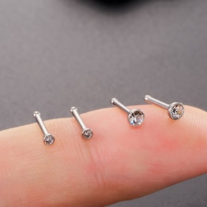 May include: Three silver nose rings with clear rhinestones. The rings are lined up on a finger.