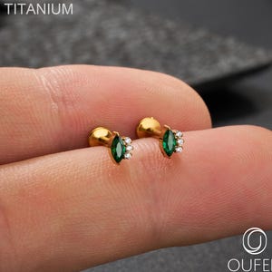 May include: Gold-coloured stud earrings featuring a marquise-cut green gemstone and small white gemstones. The word "TITANIUM" is printed in white at the top left. The brand name "OUFER BODY JEWELRY" is printed at the bottom right.