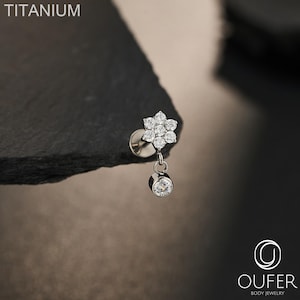 May include: A titanium body jewelry stud featuring a flower-shaped cluster of six clear round stones and a single dangling clear round stone. The text TITANIUM is visible in the upper left corner and the OUFER BODY JEWELRY logo is in the bottom right.