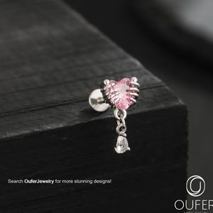 Pode incluir: Um brinco de joia corporal prateado com uma gema rosa em forma de coração e uma gema pendurada em forma de gota. O coração é emoldurado por um design prateado. O texto "Search OuferJewelry for more stunning designs!" é visível.