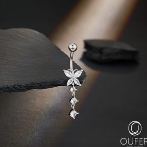May include: A silver belly button ring featuring a butterfly design with clear, sparkling accents. The butterfly is the focal point, with three dangling clear gems below. The jewelry is displayed on a dark, textured surface.