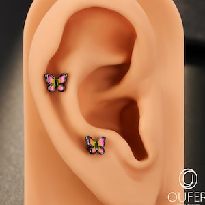 Può includere: Due orecchini a forma di farfalla con dettagli rosa, gialli e neri sono esposti su un modello di orecchio color beige. Gli orecchini sono piccoli e delicati, con un contorno nero. Lo sfondo è grigio scuro.