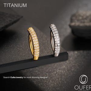 Op de afbeelding: Twee gebogen titanium body sieraden ringen, één goudkleurig en één zilverkleurig, elk bezet met kleine, sprankelende stenen. Het woord "TITANIUM" staat bovenaan. De tekst "Search OuferJewelry for more stunning designs!" staat onderaan.