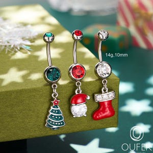 Puede incluir: Tres piercings para el ombligo con temática navideña. Cada anillo tiene una barra curva con una joya en la parte superior. Uno presenta un dije de árbol de Navidad verde, otro un gorro de Papá Noel y el último un calcetín rojo. El texto "14g, 10mm" es visible.