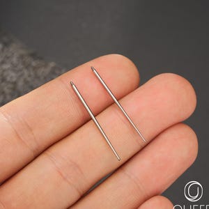 May include: Two silver-colored needles are held in a hand. The needles are thin and have a small, rounded tip. The needles are for body jewelry.
