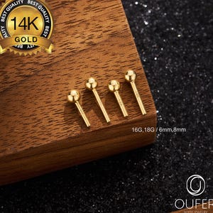Puede incluir: Cuatro pendientes de botón dorados con extremos de bola se muestran sobre una superficie de madera. Los pendientes están etiquetados como 16G y 18G, con una longitud de 6 mm y 8 mm. Un sello dorado dice "14K Gold". La marca "OUFER BODY JEWELRY" también es visible.