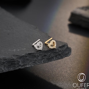 May include: Two heart-shaped nose studs, one silver and one gold, each encrusted with small, sparkling stones. The jewelry is displayed on a dark, textured surface, showcasing the delicate design and craftsmanship of the body jewelry.
