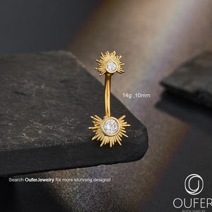 Puede incluir: Un piercing para el ombligo de color dorado con un diseño de sol. El anillo presenta dos soles, cada uno con una gema redonda y transparente en el centro. El texto "14g, 10mm" es visible. También aparece el texto "Search OuferJewelry for more stunning designs!"