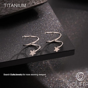 Może przedstawiać: Para srebrnych nausznic w kolorze srebrnym z motywem gwiazdy. Nausznice wykonane są z tytanu i mają spiralny kształt. Tekst "TITANIUM" jest widoczny u góry zdjęcia. Widoczny jest również tekst "Search OuferJewelry for more stunning designs!".