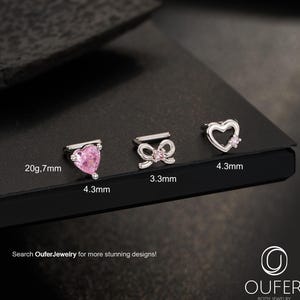 Könnte beinhalten: Drei Körperschmuckstücke werden gezeigt. Das erste ist ein herzförmiger rosa Stecker, das zweite ist ein schleifenförmiger Stecker mit einem rosa Edelstein und das dritte ist ein herzförmiger Stecker mit einem rosa Edelstein. Der Text "Search OuferJewelry for more stunning designs!" ist sichtbar.