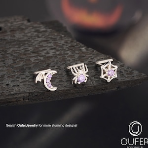Op de afbeelding: Drie zilveren lichaamsjuwelen met Halloween-thema's: een halve maan met paarse edelstenen, een spin en een spinnenweb, elk met een paarse edelsteen. De sieraden worden getoond op een donker, getextureerd oppervlak. De tekst "Search OuferJewelry for more stunning designs!" is zichtbaar.