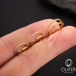 May include: Three gold-coloured double-band rings, each with a row of small, clear stones on the top band. The rings are displayed on a finger against a black backdrop. The rings vary in size.