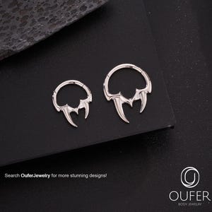 Op de afbeelding: Twee zilverkleurige lichaamsjuwelen met een tandontwerp. De sieraden zijn cirkelvormig met drie puntige tanden die zich van onderen uitstrekken. De tekst "Search OuferJewelry for more stunning designs!" is zichtbaar.