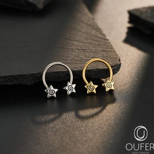 Peut inclure: Deux bijoux de corps en forme de fer à cheval. L'un est argenté, l'autre doré. Chacun a deux breloques en forme d'étoile avec des motifs en spirale. Les bijoux sont présentés sur une surface sombre et texturée. La marque "OUFER BODY JEWELRY" est visible.