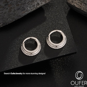 May include: Two silver-colored hoop earrings with a braided design. The earrings are circular with a hinged closure. The image includes the text "Search OuferJewelry for more stunning designs!" and the Oufer Body Jewelry logo.