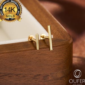 May include: A pair of gold-colored, straight bar stud earrings. The earrings are displayed on a wooden surface, with a gold seal that reads "14K GOLD" in the upper left corner. The earrings are part of the "OUFER BODY JEWELRY" collection.