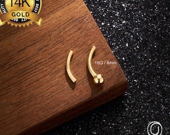 May include: Two gold-colored, curved body jewelry pieces, one with a small ball at the end, are displayed on a wooden surface. The text "14K GOLD" is visible, along with the size "16G / 8mm". The brand name "OUFER BODY JEWELRY" is also shown.