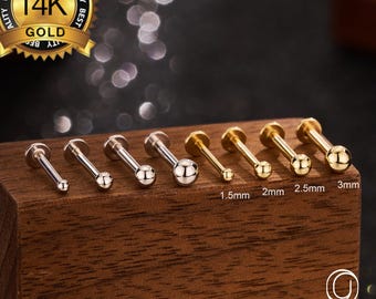 以下が含まれることがあります： 1.5mm、2mm、2.5mm、3mmのサイズの14Kゴールドとシルバーのボディジュエリースタッドのコレクション。スタッドは木製のブロックに表示され、「14K Gold」と書かれた金のシールが付いています。「OUFER BODY JEWELRY」のブランド名も表示されています。