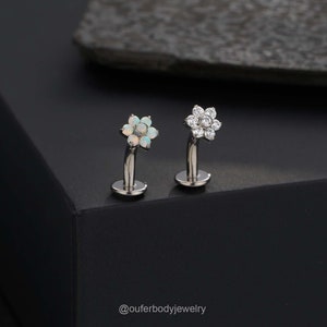 May include: Two silver flower-shaped body jewelry pieces with clear and opal stones. The jewelry is on a black surface. @ouferbodyjewelry