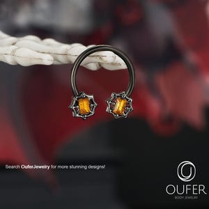 May include: A black horseshoe-shaped body jewelry with two orange-yellow rectangular gems. The gems are set in a starburst design. The jewelry is displayed against a blurred background with the text "Search OuferJewelry for more stunning designs!"