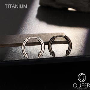 May include: Two circular horseshoe-shaped body jewelry pieces with pointed spike ends. One piece is silver-toned and the other is dark metallic gray. The text TITANIUM is visible in the upper left corner and the OUFER BODY JEWELRY logo is in the bottom right.