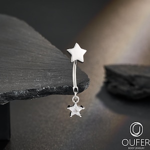 Op de afbeelding: Een zilverkleurige navelpiercing met een sterontwerp. De gebogen staaf heeft een effen ster aan de bovenkant en een bungelende ster met een heldere edelsteen aan de onderkant. De sieraden worden gepresenteerd tegen een donkere achtergrond.