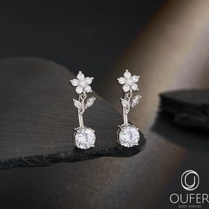 May include: A pair of silver-toned belly button rings. Each ring features a flower design at the top with a clear, round gemstone at the bottom. The rings are adorned with small, clear gemstones and leaf accents. The brand name "OUFER BODY JEWELRY" is visible.