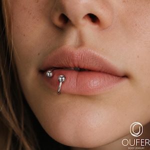 Puede incluir: Un aro labial circular de color plateado con dos cuentas esféricas pulidas en cada extremo. La joya está colocada en el labio inferior. En la esquina inferior derecha aparece el texto blanco OUFER BODY JEWELRY con un icono de logo circular.