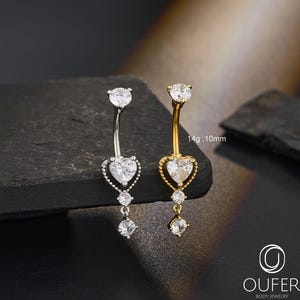 May include: Two belly button rings, one silver-toned and one gold-toned, each featuring a heart-shaped center stone and dangling crystals. The gold ring has a heart-shaped frame. The text "14g, 10mm" is visible. The logo for Oufer Body Jewelry is in the bottom right.