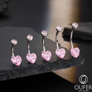 May include: Five belly button rings with a curved barbell design. Each ring features a pink heart-shaped gem and a small, round gem. The jewellery is displayed on a dark, textured surface.