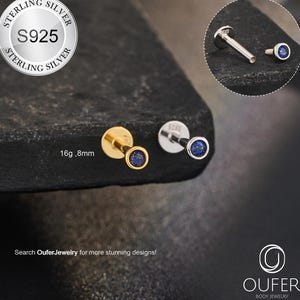 May include: A close-up of two stud earrings, one gold and one silver, each with a small, round, dark blue gemstone. The earrings are displayed on a dark, textured surface. The text "STERLING SILVER S925" is visible.