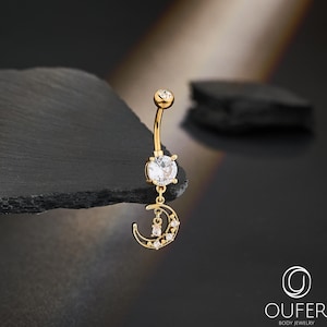 May include: A gold-colored belly button ring with a curved barbell design. The ring features a large, clear gemstone at the center and a dangling crescent moon charm with smaller gemstones. The jewelry is set against a dark background.