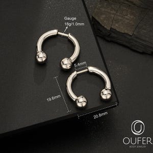 May include: Two silver-colored circular horseshoe-shaped body jewelry pieces with ball ends. The gauge is 18g/1.0mm. One piece measures 5.4mm by 19.6mm, and the other measures 20.8mm. The jewelry is displayed with technical measurement labels.