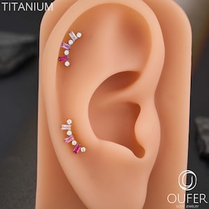 Puede incluir: Pendientes de titanio con un diseño curvo. Los pendientes presentan una combinación de piedras preciosas claras y rosas con corte baguette y corte redondo. Los pendientes se muestran en un modelo de oreja color carne. La palabra "TITANIUM" está impresa en la parte superior.
