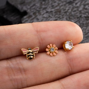 May include: Three gold-toned body jewelry pieces: a bee with black and pink enamel, a pink flower with a clear rhinestone center, and a clear round rhinestone.