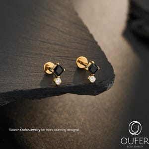 Puede incluir: Un par de pendientes de oro con una piedra negra de corte cuadrado y una piedra transparente redonda más pequeña debajo. Los pendientes tienen un diseño de cierre plano. El texto OuferJewelry es visible en la esquina inferior derecha.