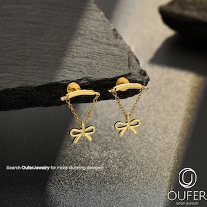 May include: A pair of gold-toned earrings featuring a curved bar post connected by two delicate chains to a small bow charm. The earrings are displayed with the text Search OuferJewelry for more stunning designs and the Oufer Body Jewelry logo.